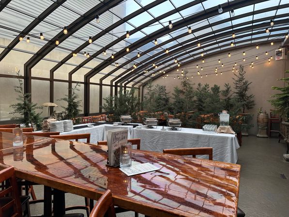 Glass-roofed covered patio restaurant with arched metal beams and warm string lights, glossy wooden communal table in foreground, buffet setup with chafing dishes and water dispenser, evergreen plants and wooden chairs in a cozy courtyard setting
