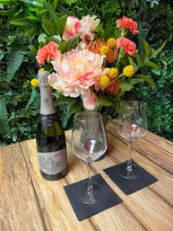 Cheery floral arrangement of pink peony, coral carnations, orange pincushion protea and yellow billy balls beside a bottle of cava and two empty wine glasses on a wooden patio table against a green living wall