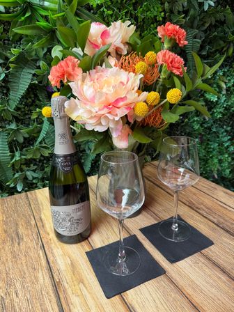 Cheery floral arrangement of pink peony, coral carnations, orange pincushion protea and yellow billy balls beside a bottle of cava and two empty wine glasses on a wooden patio table against a green living wall