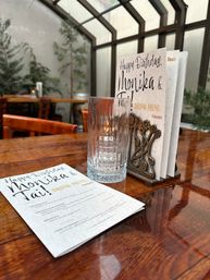 Glass tumbler and folded birthday drink menu in an ornate holder on a polished wooden table at a sunlit indoor garden-style restaurant, with leafy plants, wooden chairs and skylight windows.