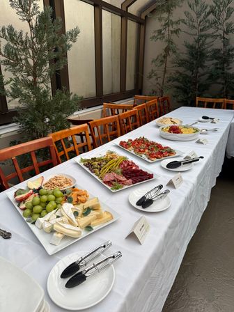 Outdoor patio buffet on a white-linen table with cheese and fruit platter, charcuterie, bruschetta and dips, wooden chairs and tall potted evergreens in a courtyard setting.