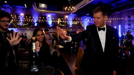 Man in tuxedo reaching for a playful high-five with two guests over a champagne bottle in a purple-lit industrial event space decorated with string lights and garlands.