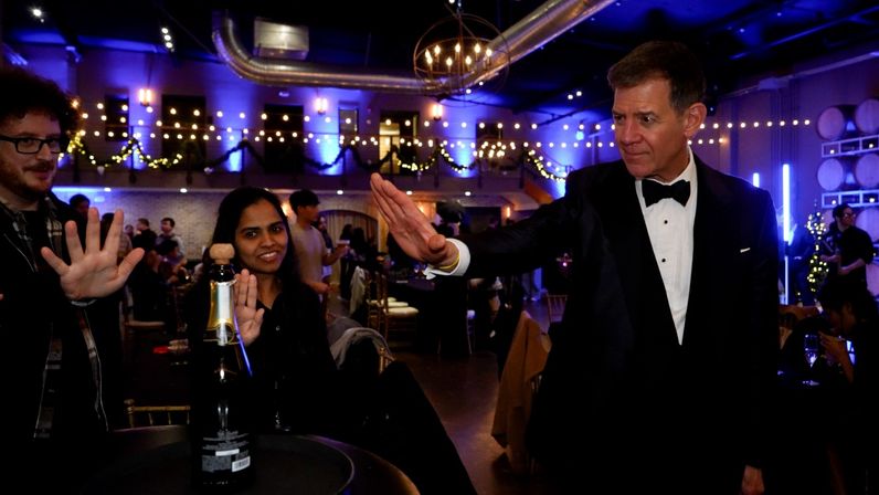 Man in tuxedo reaching for a playful high-five with two guests over a champagne bottle in a purple-lit industrial event space decorated with string lights and garlands.