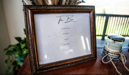 Vintage-style framed 'The Bar' sign listing cocktails and wine on a wooden table next to a twine-tied jar candle, with potted greenery and a sunlit balcony in the background.