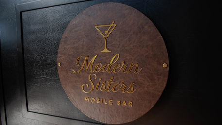 Round brown leather-look plaque with gold martini-glass icon and elegant gold lettering that reads mobile bar, mounted on a dark wood wall, sleek modern bar sign.