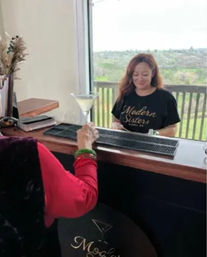 Customer at a wooden bar counter receives a martini from a smiling bartender wearing a black branded T-shirt, with a bar mat on the counter and a green landscape visible through a large window/balcony.