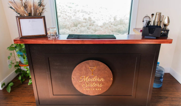 Indoor portable wooden bar with reddish countertop and round logo plaque, cocktail shakers and bartending tools on the right, dried floral arrangement and potted plant on the left, water jug nearby and a large window framing a hilly landscape.