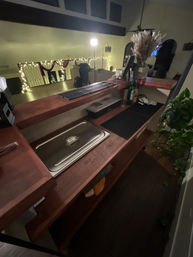 Cozy loft home bar with wooden counter, stainless serving pan, bottles and a vase of pampas grass, overlooking a living room balcony draped in warm string lights