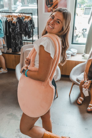 Smiling woman in a boutique trying on an oversized round pink faux-fur tote over a white tee and shorts, standing by clothing racks and seating.