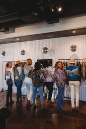 Group of women in a trendy boutique trying on sparkly fringe jackets and denim, posing by clothing racks and disco ball decor with cowboy hats and boots for a playful Western vibe.
