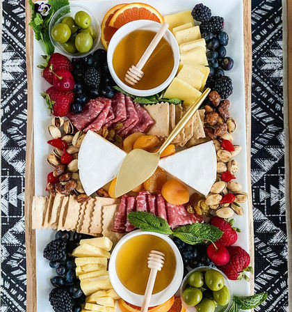 Vibrant charcuterie board on a wooden tray with brie wedges, sliced cheeses, salami, green olives, fresh berries, dried apricots, nuts, crackers and two honey bowls