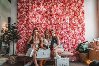 Three friends smiling on a bench with shopping bags in a trendy boutique, posing in front of a pink rose flower wall and neon sign that reads 'you're like, really pretty', flanked by potted plants.