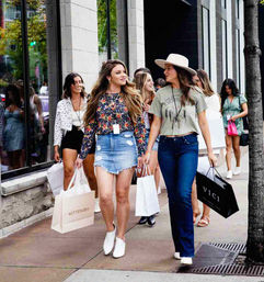 Group of young women shopping on a downtown urban sidewalk, two in the foreground—one in a floral top and denim mini skirt and the other in a wide‑brim hat and jeans—carrying boutique shopping bags and chatting in front of storefront windows.