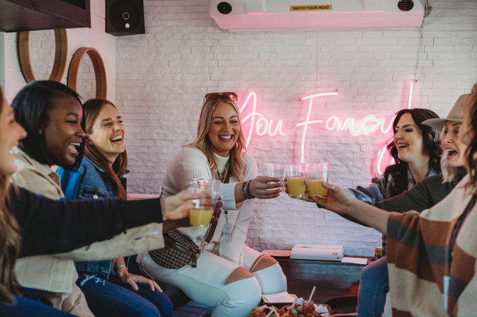 Group of smiling women clinking mimosas at a trendy indoor brunch, seated by a white brick wall with a pink neon sign.