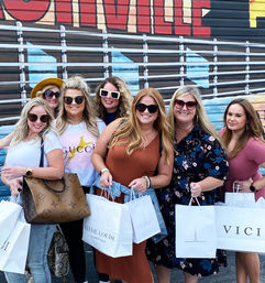 Seven smiling women in sunglasses and casual outfits holding boutique shopping bags, posing as friends in front of a colorful urban mural on a sunny day.