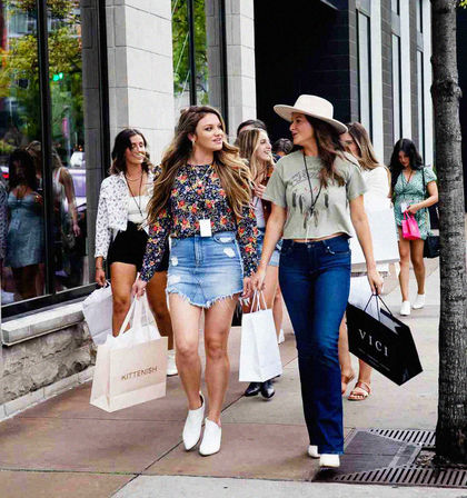 Group of young women shopping on a downtown urban sidewalk, two in the foreground—one in a floral top and denim mini skirt and the other in a wide‑brim hat and jeans—carrying boutique shopping bags and chatting in front of storefront windows.