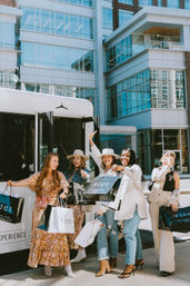 Five stylish women laughing with shopping bags on a downtown sidewalk beside a white boutique shuttle, modern glass buildings behind