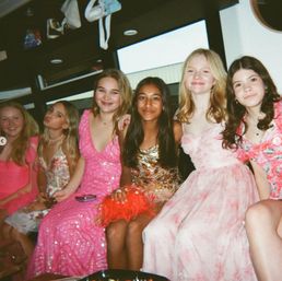 Six teenage friends in colorful party dresses seated together inside a party bus, smiling and posing for a lively group photo during a celebration.