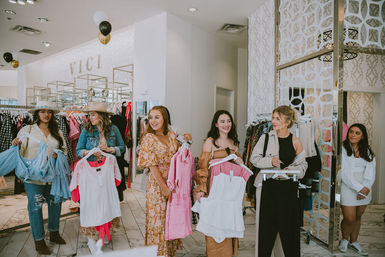Group of women shopping in a bright fashion boutique, smiling as they hold colorful dresses and tops on hangers amid clothing racks and stylish patterned walls.