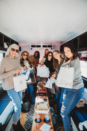 Group of women smiling inside a cozy party bus, holding shopping bags around a wooden table with charcuterie, snacks and drinks — daytime shopping trip and mobile brunch.