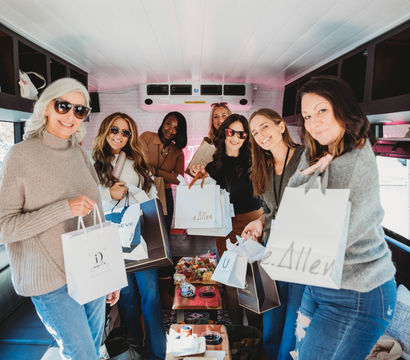 Group of women smiling inside a cozy party bus, holding shopping bags around a wooden table with charcuterie, snacks and drinks — daytime shopping trip and mobile brunch.