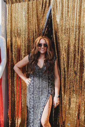 Smiling woman in round sunglasses wearing a beaded black star-pattern dress with a high slit, posing in front of a shimmering gold sequin event backdrop.