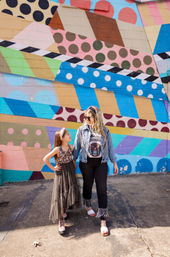 Woman and young girl holding hands and smiling while walking in front of a colorful geometric and polka-dot mural on an urban sidewalk.