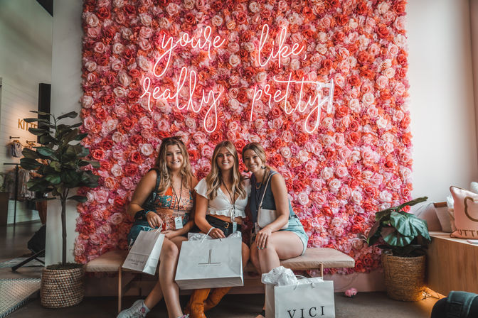 Three friends smiling on a bench with shopping bags in a trendy boutique, posing in front of a pink rose flower wall and neon sign that reads 'you're like, really pretty', flanked by potted plants.