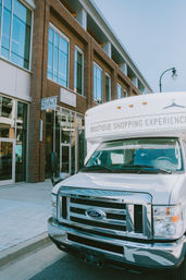 White mobile boutique van parked on a downtown shopping street in front of a red brick retail building with large glass windows and a boutique sign