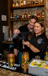 Bartender pours a bright blue cocktail from a pitcher into a jigger behind a brick-walled bar as a man watches, with liquor bottles on shelves, colorful painted cups, and a fruit garnish tray on the counter.