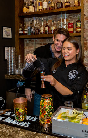 Bartender pours a bright blue cocktail from a pitcher into a jigger behind a brick-walled bar as a man watches, with liquor bottles on shelves, colorful painted cups, and a fruit garnish tray on the counter.