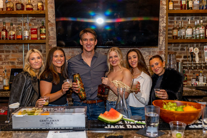 Six friends smiling together at a rustic brick-walled bar, holding cocktails and bottles behind a counter with a watermelon slice, fruit bowl and glasses, with liquor shelves and beer taps in the background.
