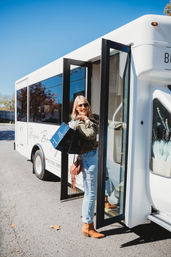 Smiling woman in sunglasses stepping off a white boutique shopping shuttle on a sunny day, holding blue and black shopping bags and wearing light jeans, a fringe crossbody bag, and brown ankle boots.