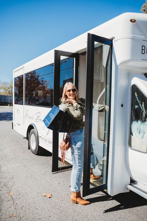 Smiling woman in sunglasses stepping off a white boutique shopping shuttle on a sunny day, holding blue and black shopping bags and wearing light jeans, a fringe crossbody bag, and brown ankle boots.
