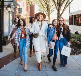 Smiling group of women friends strolling a tree-lined downtown sidewalk in fall, wearing casual autumn outfits, boots and hats, carrying shopping bags and event lanyards.