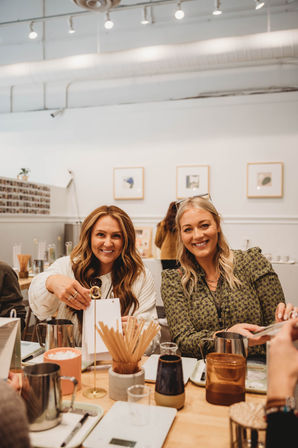 Two smiling women enjoying a coffee tasting at a bright modern café, with milk pitchers, scales, wooden stirrers and cups on a wooden table beneath minimalist white walls and framed art