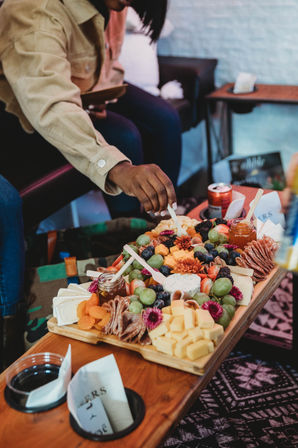 Hand reaching for a colorful charcuterie board with cubed and soft cheeses, grapes, berries, dried apricots, folded cured meats, jam jars and edible flowers on a wooden serving board in a cozy lounge setting.