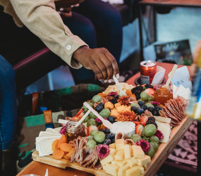 Hand reaching for a colorful charcuterie board with cubed and soft cheeses, grapes, berries, dried apricots, folded cured meats, jam jars and edible flowers on a wooden serving board in a cozy lounge setting.