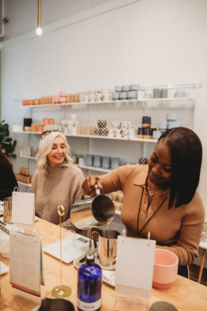 Two smiling women in a bright craft studio pouring wax during a hands-on candle-making workshop, with scales, metal pitchers and shelves of colorful ceramic containers in the background.