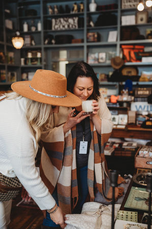 Two women shopping in a cozy downtown boutique—one in a tan wide‑brim hat—smelling a scented candle over a display of blankets and artisanal home decor on wooden shelves