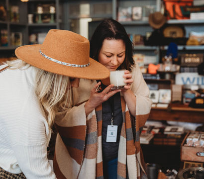 Two women shopping in a cozy downtown boutique—one in a tan wide‑brim hat—smelling a scented candle over a display of blankets and artisanal home decor on wooden shelves