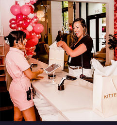 Smiling customer receives a white shopping bag from a cheerful sales associate at a boutique checkout counter with pink balloon decorations