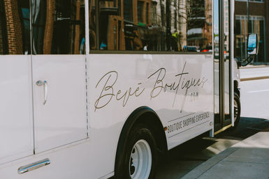 White boutique shuttle bus parked on a downtown street, with stylish cursive logo and 'boutique shopping experience' text along the side