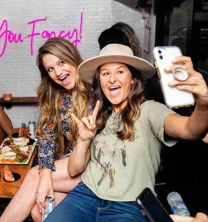 Two friends taking a selfie at an indoor casual brunch — woman with a wide-brim hat flashes a hand sign while the other in a floral dress holds a canned drink, with an appetizer platter and neon "You Fancy!" sign in the background.