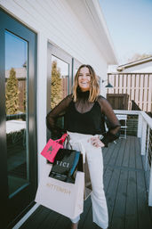 Smiling woman on a modern backyard deck holding colorful shopping bags, wearing a sheer black blouse and white high-waist pants — bright, stylish outdoor shopping moment.