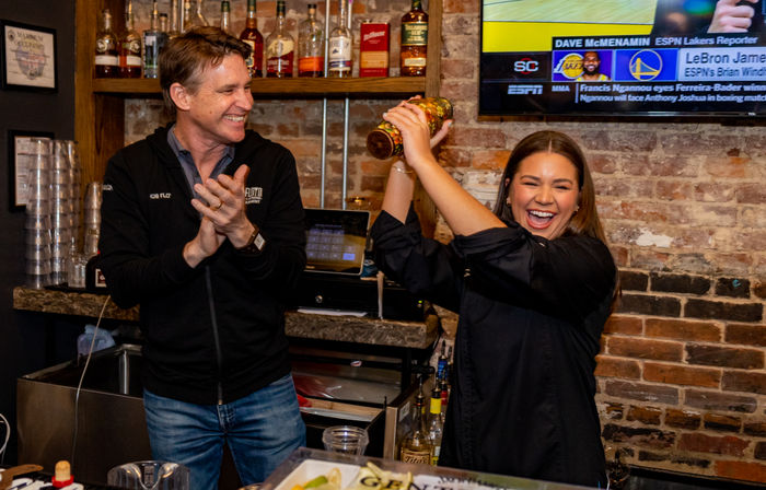 Smiling bartender energetically shaking a cocktail shaker behind a brick-walled bar as a man claps, with liquor bottles on shelves and a sports TV on the wall.