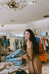 Smiling woman shopping in a bright women's boutique, folding light-wash jeans at a display table with sandals and colorful clothing racks in the background.