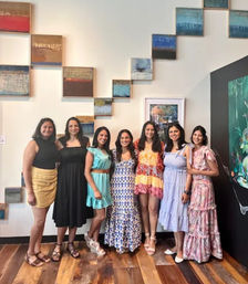 Seven women in colorful summer dresses smiling and posing inside an art gallery in front of stepped abstract wall panels and hardwood floors.