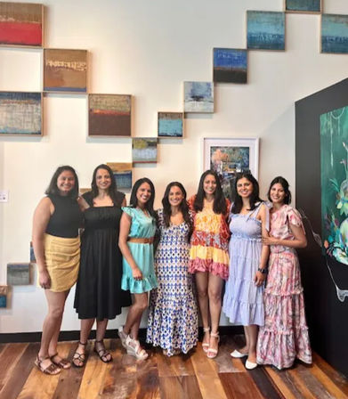 Seven women in colorful summer dresses smiling and posing inside an art gallery in front of stepped abstract wall panels and hardwood floors.