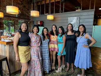 Seven women in colorful summer dresses smiling and posing inside a cozy brick-walled restaurant with wooden bar and pendant lights